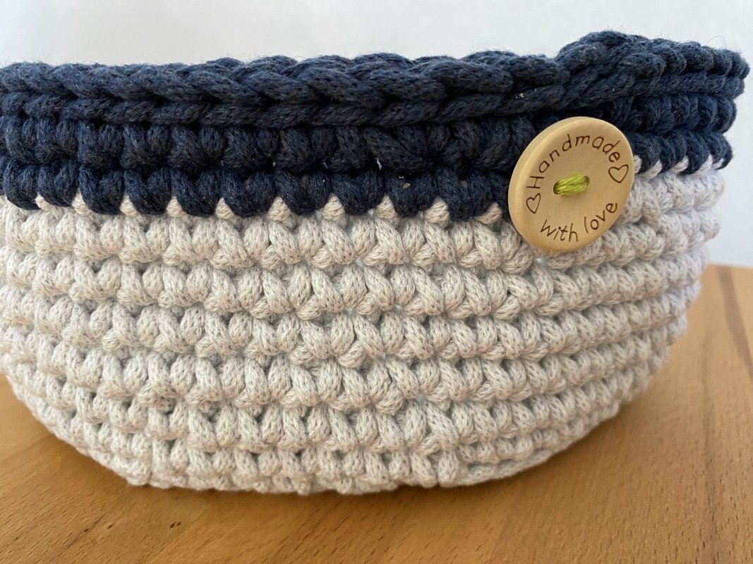 Gehäkelter Korb beige und dunkelblau mit Holzknopf 'Handmade with love' auf Holzoberfläche