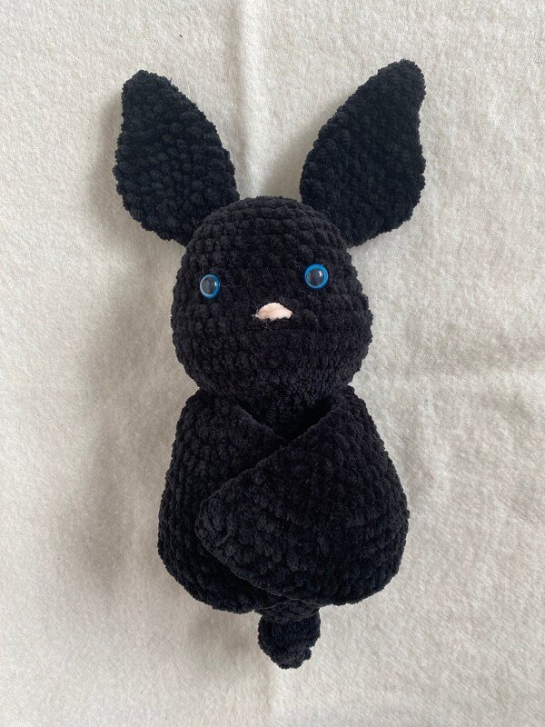 Close-up of a black crocheted bat plush toy with blue eyes and a small pink nose on a light background.