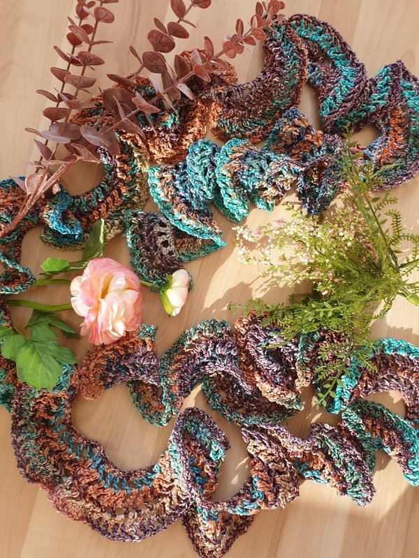Ruffled variegated crochet scarf in teal, brown, and purple on a wooden surface with flowers and botanical decor.