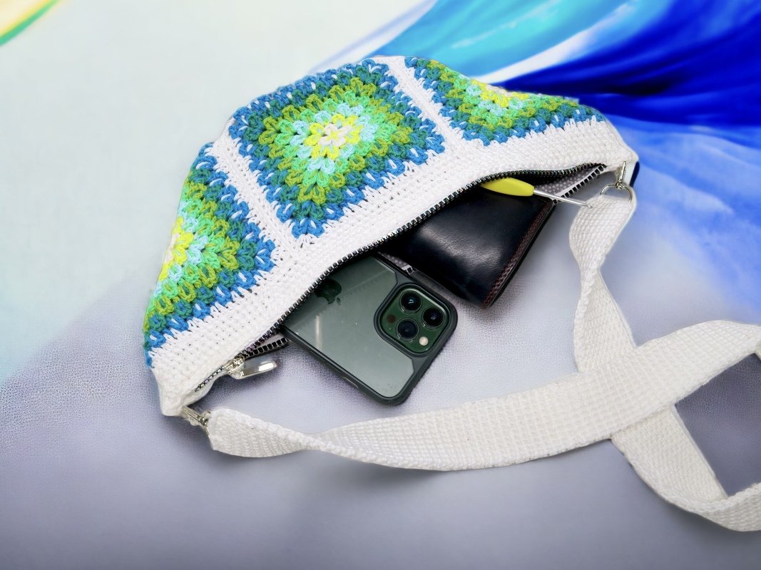 Gehäkelte Umhängetasche mit blau-grünen Granny-Square-Paneelen, weißem Gurt und offenem Reißverschluss, Smartphone und Geldbörse sichtbar