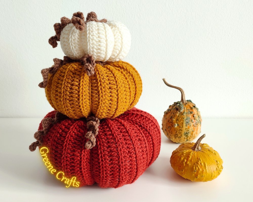 Three crocheted pumpkins stacked (cream, mustard, red) with brown stems and two small natural gourds on a white surface.