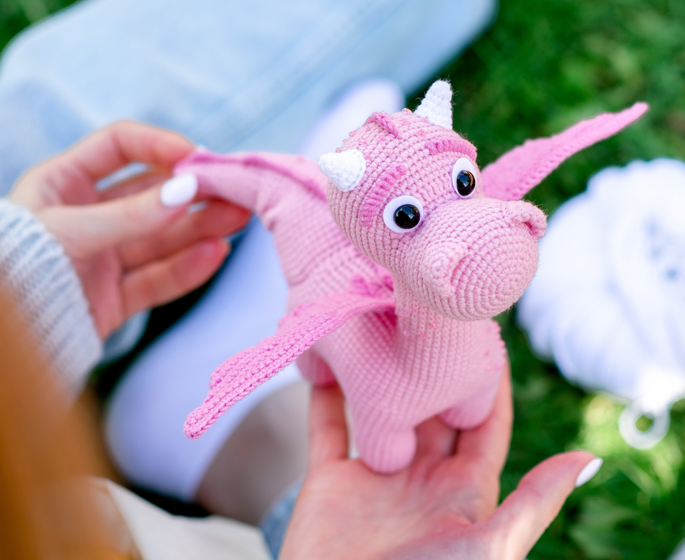 Rosa gehäkelter Amigurumi-Drache mit Hörnern, von Händen gehalten.