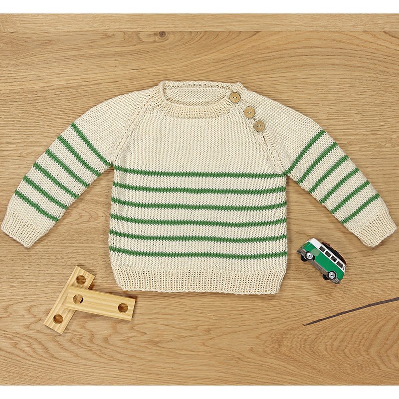Cremefarbener Kinderpullover mit grünen Streifen und drei Holzknöpfen an der Schulter, auf Holztisch liegend.