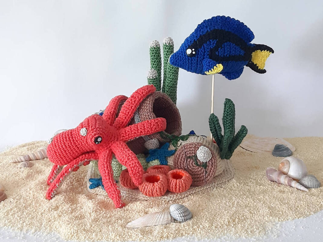 Nahaufnahme einer gehäkelten Unterwasserlandschaft mit rotem Oktopus, blauem Fisch, Korallen, Seegras und Muscheln auf Sand.