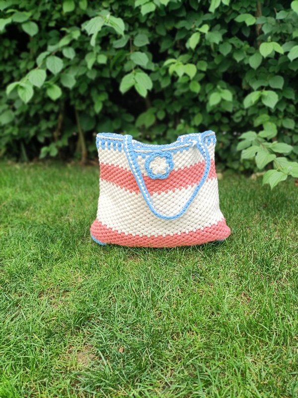 Handgehäkelter Shopper mit creme- und korallenfarbenen Streifen, hellblaue Einfassung und mittige Blüte auf Rasen
