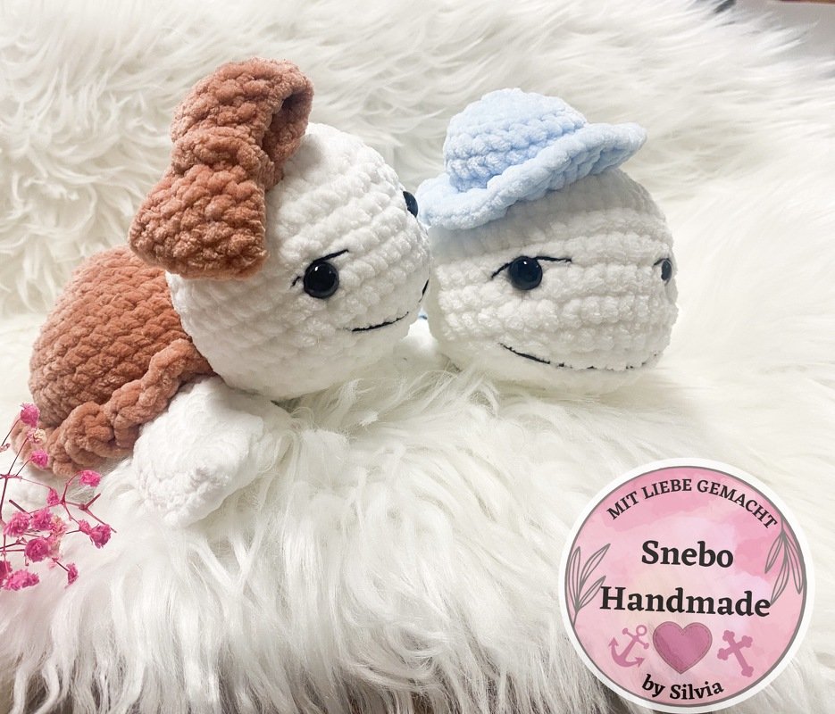 Zwei kompakte weiße Amigurumi-Wasserfiguren aus Plüschgarn, eines mit braunen Details und eines mit blauem Häubchen, auf weißem Fell