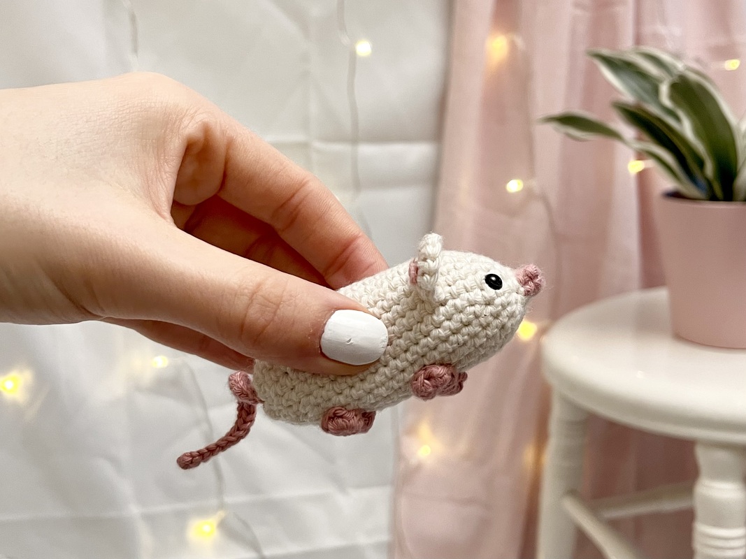 Gehäkelte cremeweiße Amigurumi‑Maus mit rosa Details, gehalten in einer Hand