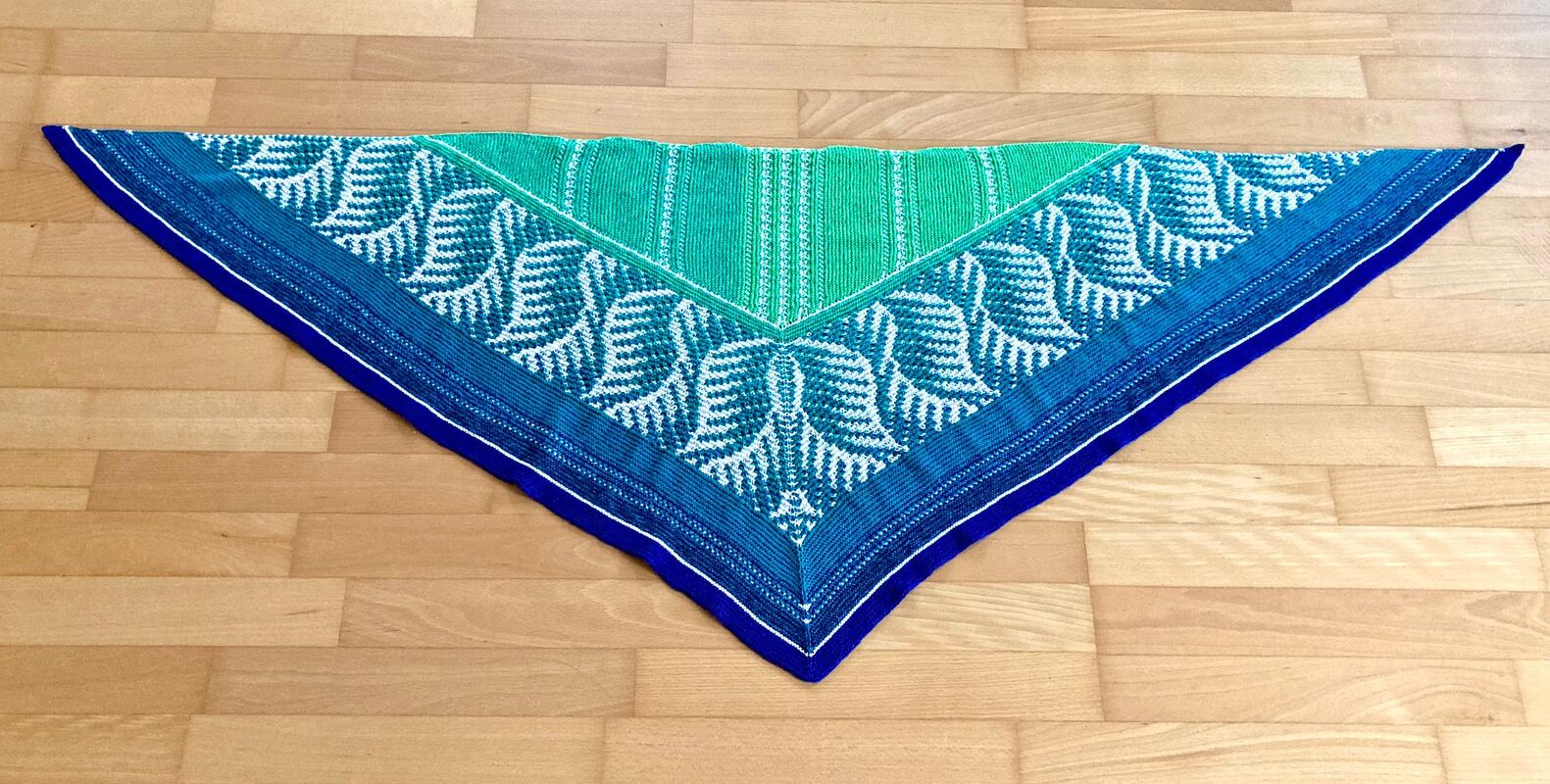 Gestricktes Dreieckstuch blau‑grün mit weißem Blattmuster auf Holzboden