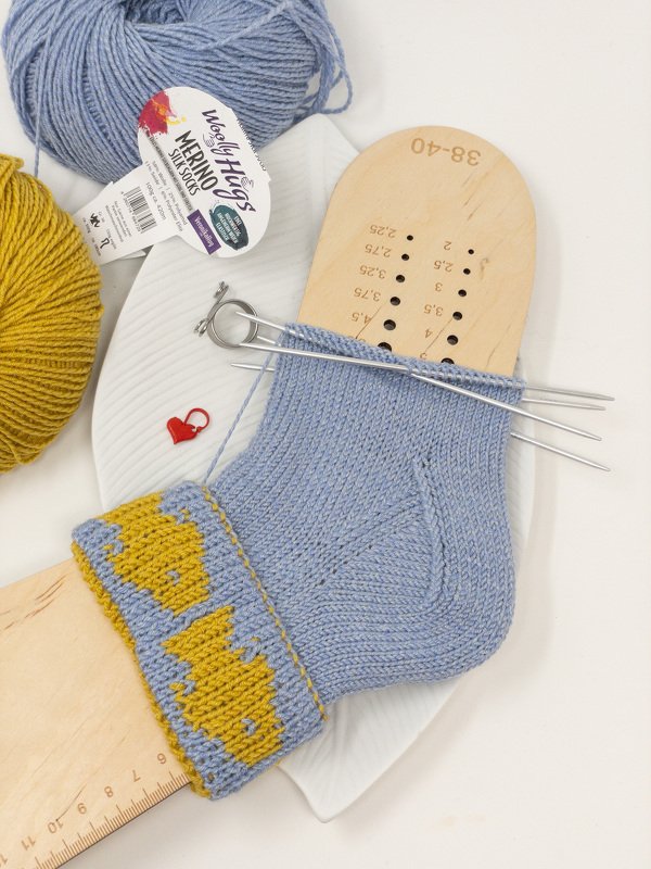 Hellblaue Socke mit gelbem Fischmuster im Bündchen auf Holz-Sockenspannform mit Nadelspiel und Wollknäueln