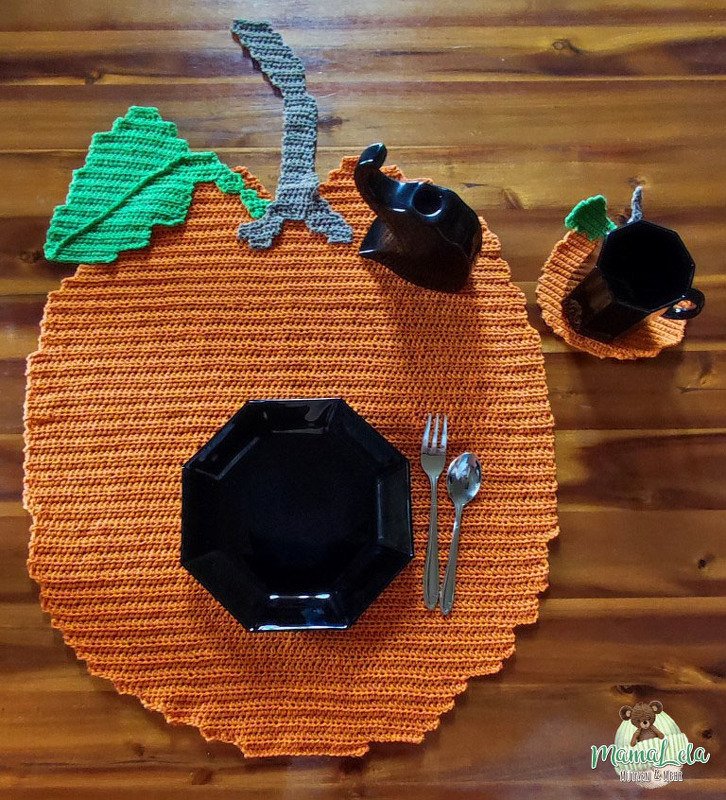 Gehäkeltes orangefarbenes Kürbis-Deckchen mit grünem Blatt und grauem Stiel, schwarzem Geschirr und Besteck auf Holztisch