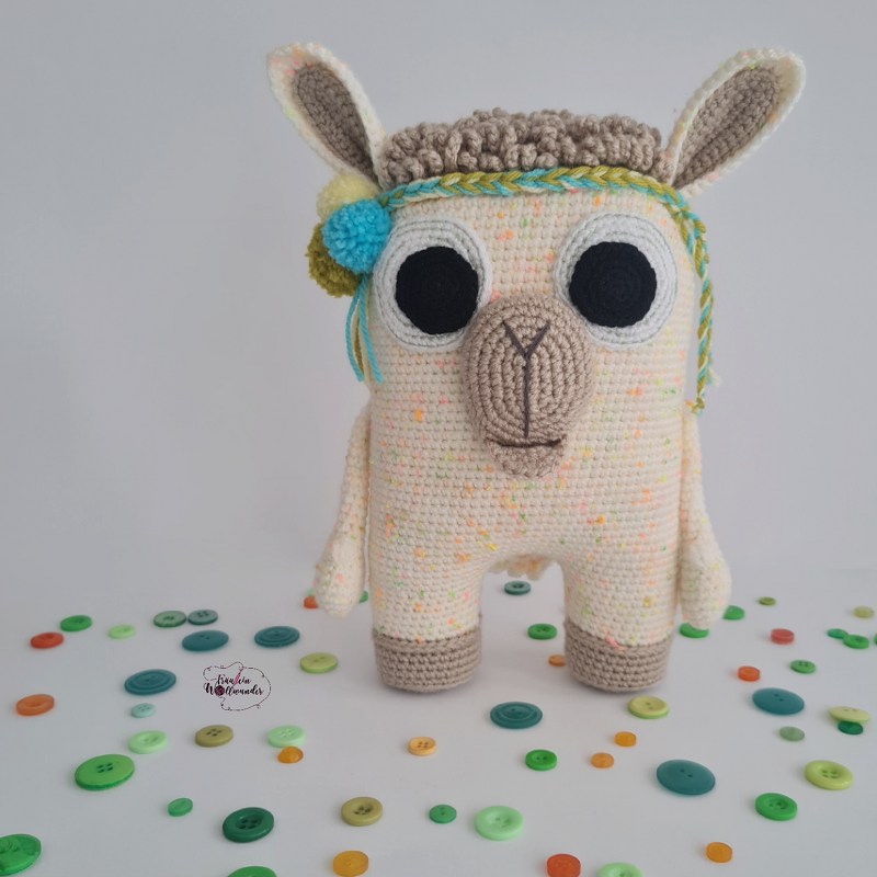 Gehäkeltes Lama-Amigurumi in Creme mit Sprenkelgarn, geflochtenem Stirnband und bunten Bommeln, umgeben von Knöpfen