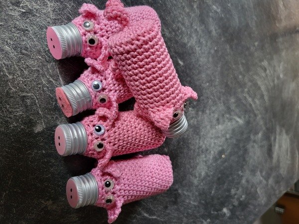 Mehrere rosa gehäkelte Überzüge mit aufgesetzten Augen und Knopf‑Nasen auf Schraubverschlüssen