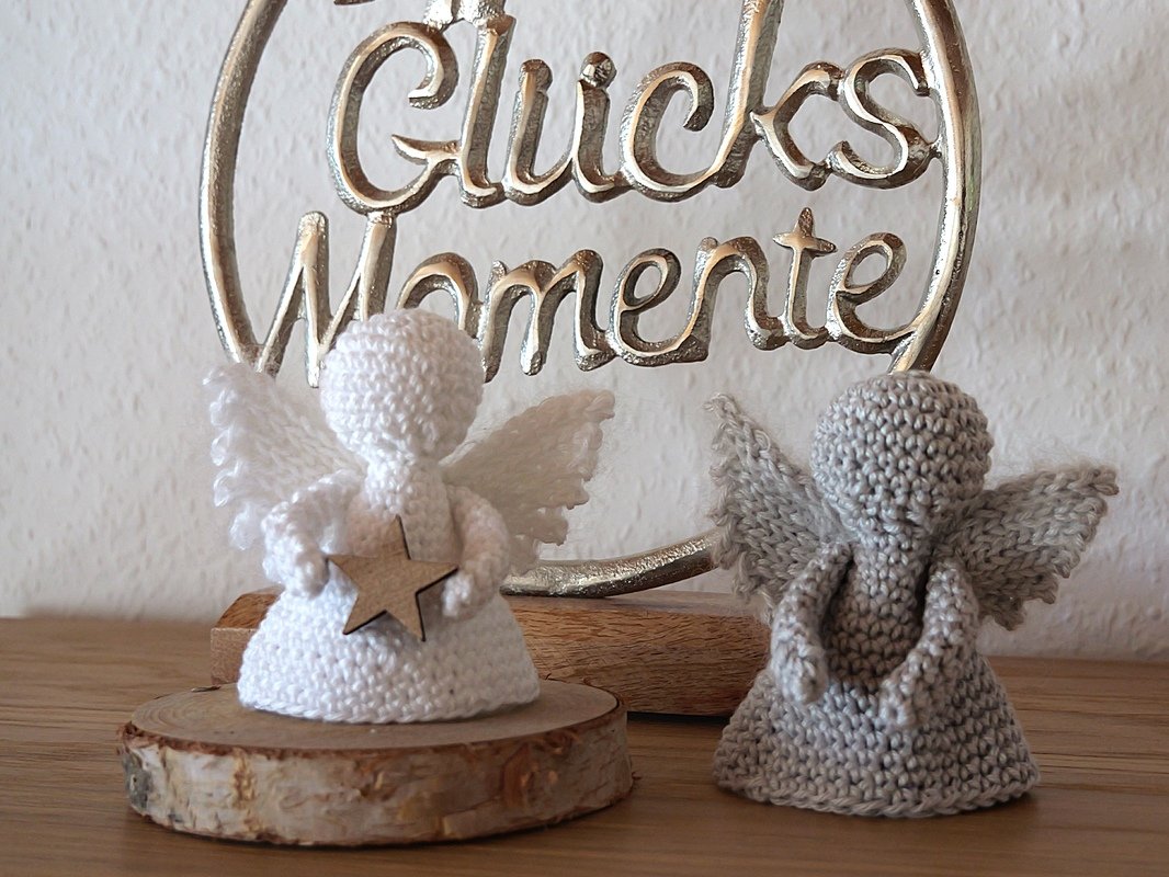 Zwei gehäkelte Amigurumi-Engel, weiß mit Holzstern auf Holzscheibe und grau, vor einem silbernen 'Glücks Momente'-Schild.