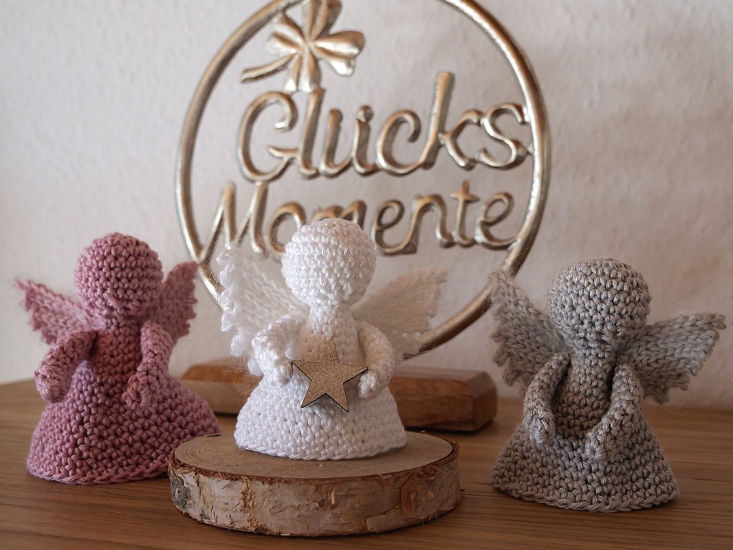 Drei gehäkelte Amigurumi-Engel (rosa, weiß, grau); weißer Engel mit Holzstern auf Baumscheibe