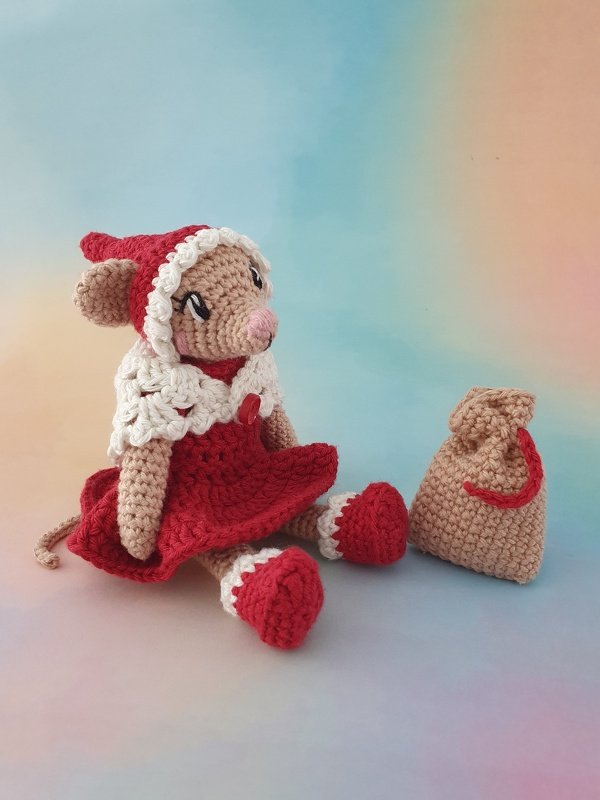 Gehäkelte Maus in rotem Kleid und Zipfelmütze sitzt neben beigem Geschenksack vor buntem Hintergrund.