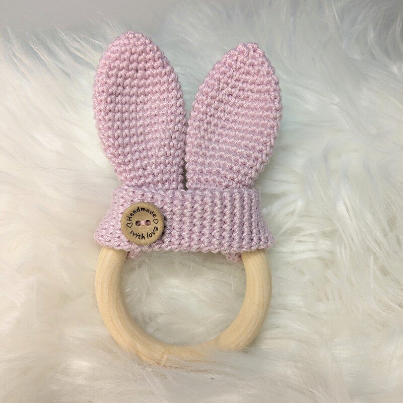 Gehäkelter Greifring mit zarten Flieder-Hasenohren und Holzring auf weißem Fell