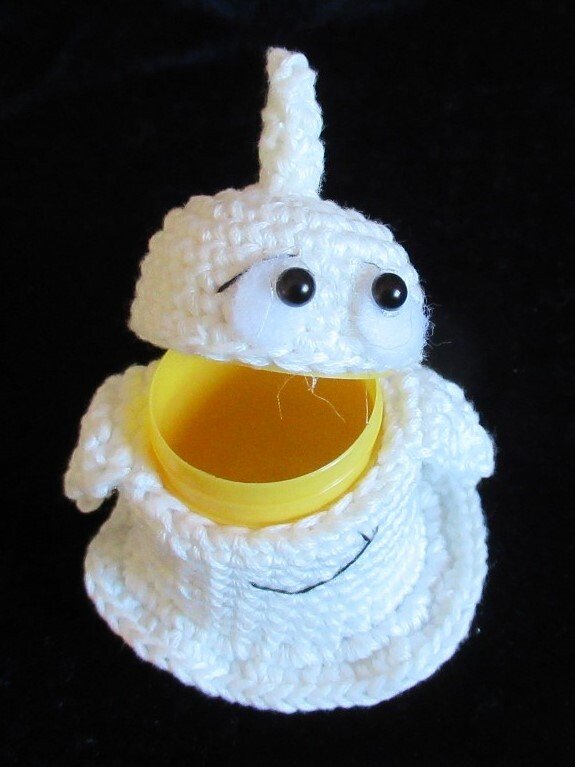 Weißer gehäkelter Geist mit geöffnetem Kopf, der eine gelbe Kunststoffdose im Inneren zeigt, vor schwarzem Hintergrund.