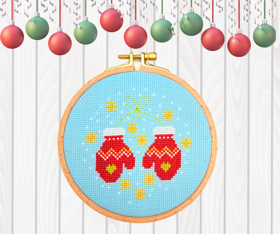 Rundes Kreuzstichbild mit roten Fäustlingen auf hellblauem Stoff im Holzstickrahmen vor weißer Holzoberfläche mit Weihnachtskugeln
