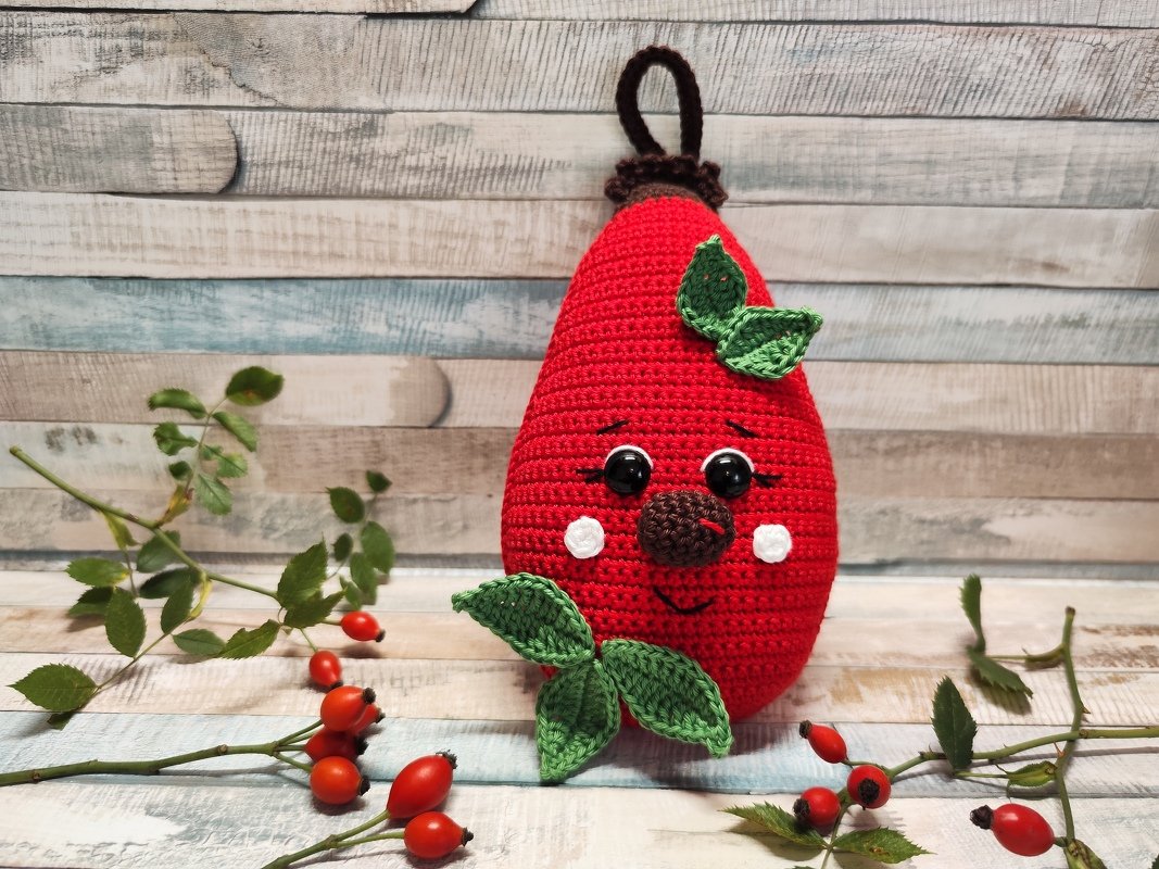 Rote, gehäkelte Hagebutte als Amigurumi mit Augen, Nase, Mund und weißen Wangenpunkten, versehen mit grünen Blättern und Aufhänger, auf Holzuntergrund neben echten Hagebuttenzweigen.