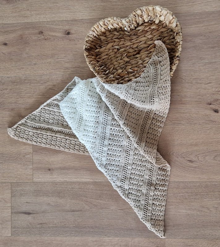 Gehäkeltes, zweifarbiges Dreieckstuch in Creme und Beige auf hellem Holzboden neben einem herzförmigen Korb.