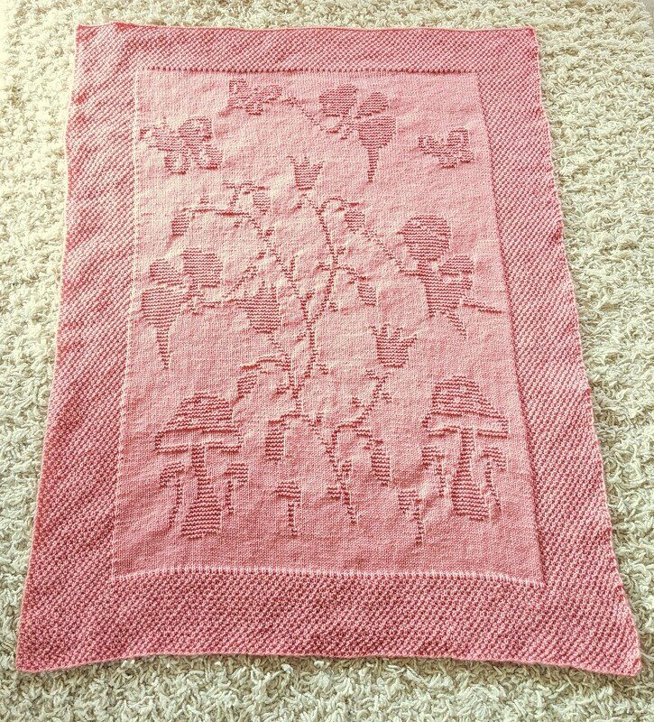 Light pink knitted baby blanket with raised mushroom, butterfly, and floral motifs on a light carpet.
