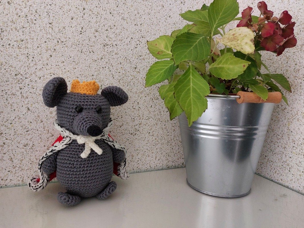 Gehäkelte graue Amigurumi-Maus mit gelber Krone und rotem Umhang neben Topfpflanze in Metallkübel