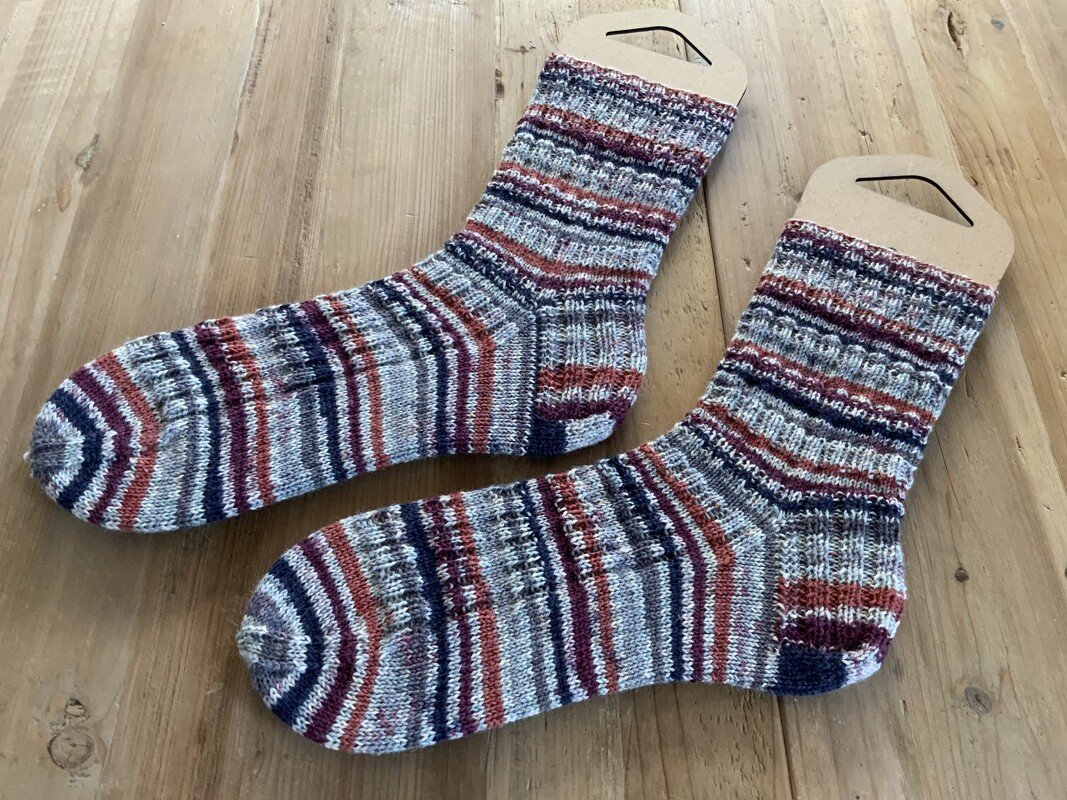 Gestrickte, gestreifte Socken in Terrakotta-, Marineblau- und Grautönen auf Holz mit Pappetiketten.