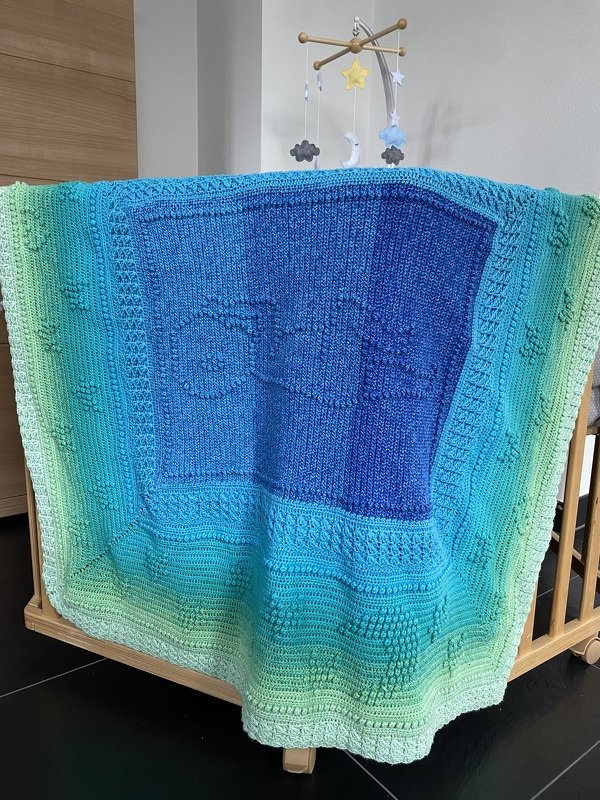 Gehäkelte Babydecke in Blau- und Grüntönen mit Wolkenmotiv über einem Babybett