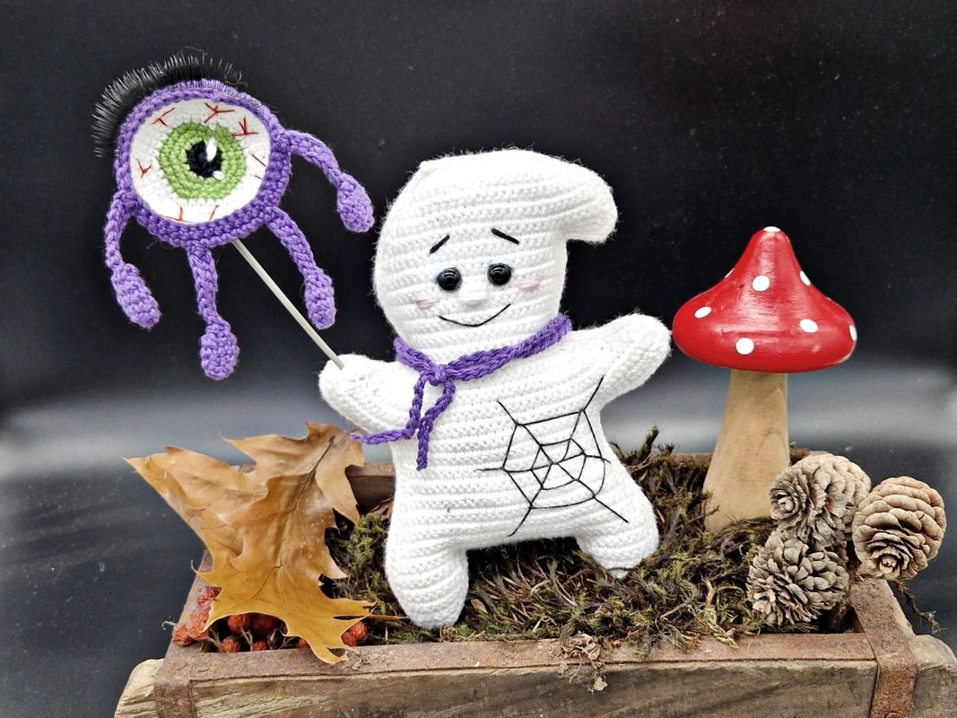 Handgehäkelter weißer Geist mit lila Schal hält lilafarbenes Monsterauge auf Stab, dekoriert mit Moos, Blättern, Tannenzapfen und Fliegenpilz.