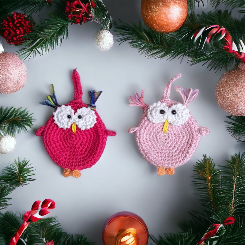 Zwei kompakte, schnell gehäkelte Eulen in Pink und Rosa mit weißen Augen und gelbem Schnabel, auf hellem Hintergrund mit Tannengrün und Weihnachtskugeln