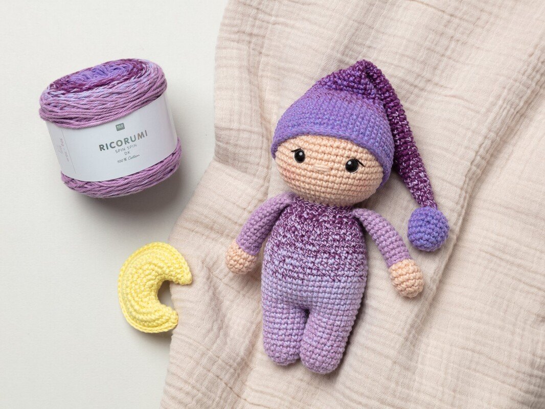 Purple-gradient amigurumi doll wearing a long pointed nightcap with pompom, next to a skein of purple yarn and a small yellow crocheted crescent moon on a beige muslin cloth.