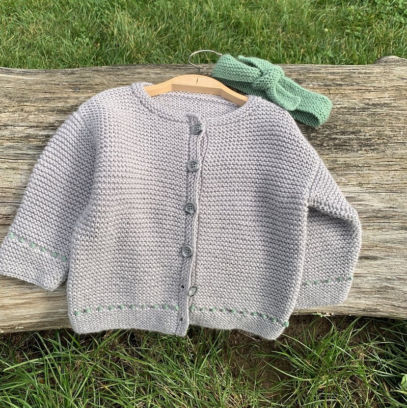 Hellgraue gestrickte Babyjacke mit fünf Knöpfen und grünem Stirnband auf Holzbügel