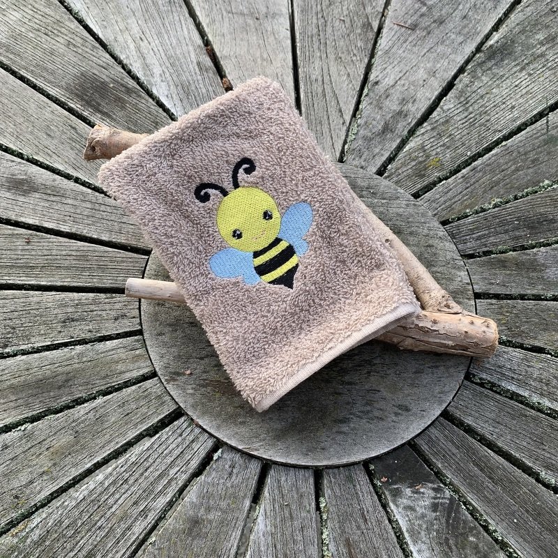 Beiges Frottiertuch mit gestickter gelb-schwarzer Biene und hellblauen Flügeln auf rundem Holzuntergrund
