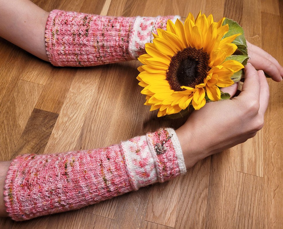 Gestrickte rosa Armstulpen an Armen, eine Hand hält eine Sonnenblume