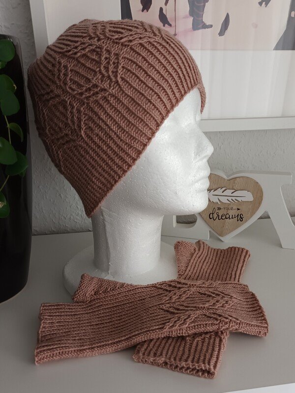 Roséfarbenes gestricktes Mützen- und Stulpen-Set mit Zopfmuster; Mütze auf Schaufensterkopf, Stulpen liegend.