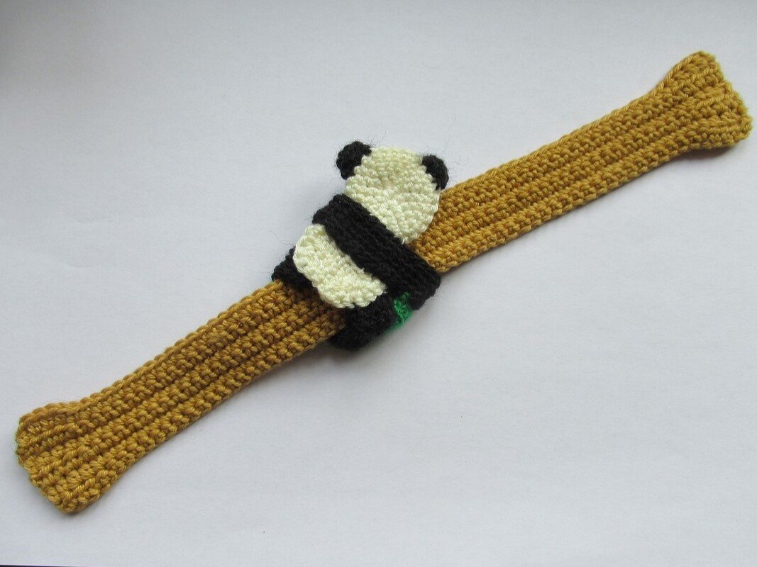 Gehäkeltes ockerfarbenes Lesezeichen mit einer kleinen schwarz-weißen Panda-Applikation, die das Band umschließt.