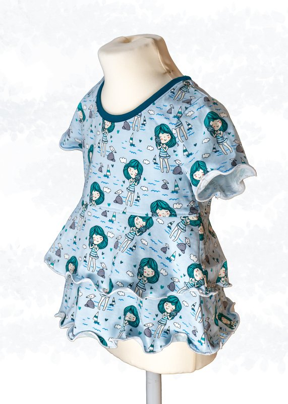 Hellblaues Kinder-Tunika-Top mit türkisfarbenem Mädchenmotiv, zweistufigen Volants und dunklem Halsbündchen auf einer Schaufensterpuppe