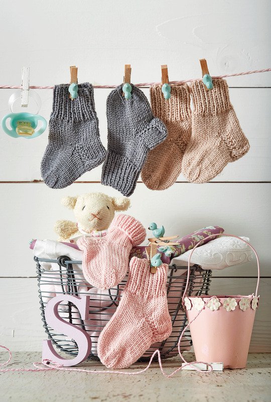 Gestrickte Babysocken in Grau und Rosa an einer Leine, darunter Drahtkorb mit Kuscheltier und weiteren Socken