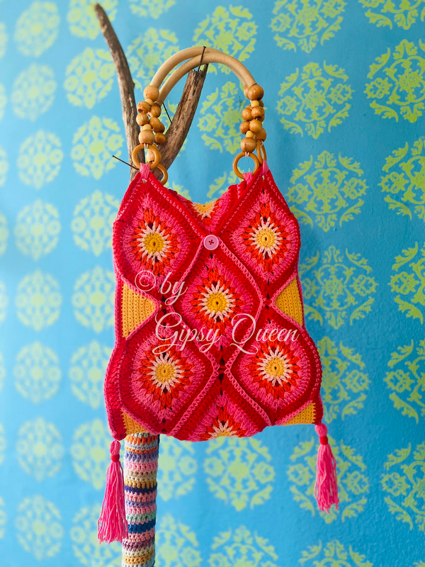 Gehäkelte Umhängetasche mit pink-orangen Granny-Diamanten, Holzgriffen und Quasten vor blau gemustertem Hintergrund