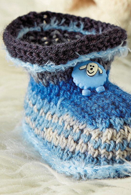 Ein gehäkelter Babyschuh in Blau-, Grau- und Weißtönen mit einem hellblauen Schafknopf auf einem flauschigen weißen Untergrund.