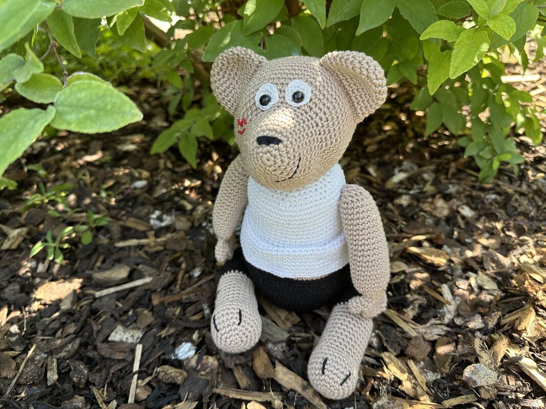 Beiger gehäkelter Teddy mit rotem Herz auf der Wange und weißer Weste, sitzend im Freien