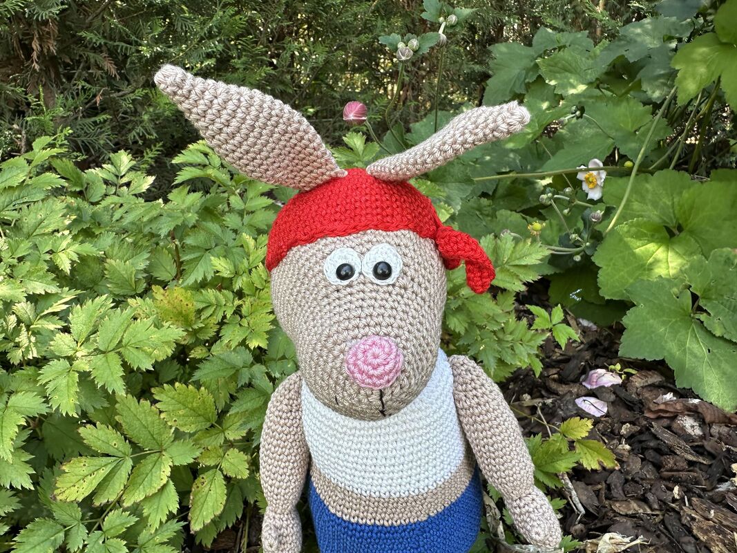 Gehäkelter Amigurumi-Hase mit langen Ohren, rotem Kopftuch, großen Augen, rosa Nase, weißem Oberteil und blauer Hose im Grünen