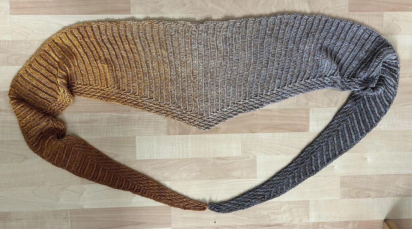 Crocheted triangle shawl with ribbed texture and a color gradient from golden brown to grey, laid flat on a wooden floor.