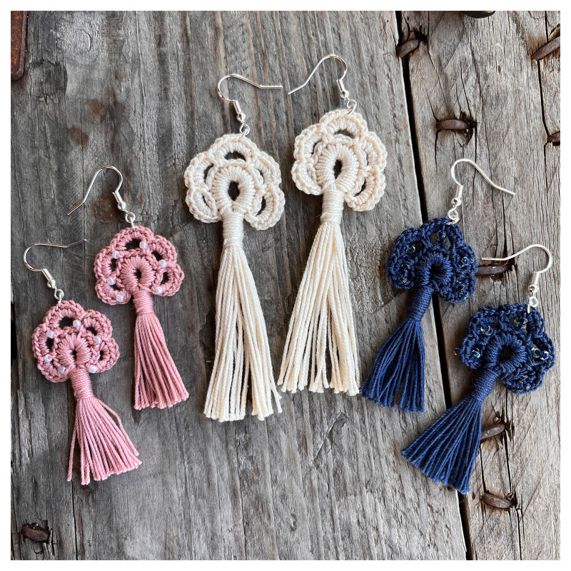 Nahaufnahme von drei Paaren gehäkelter Quasten-Ohrringe in Rosa, Creme und Marineblau auf rustikalem Holzhintergrund.