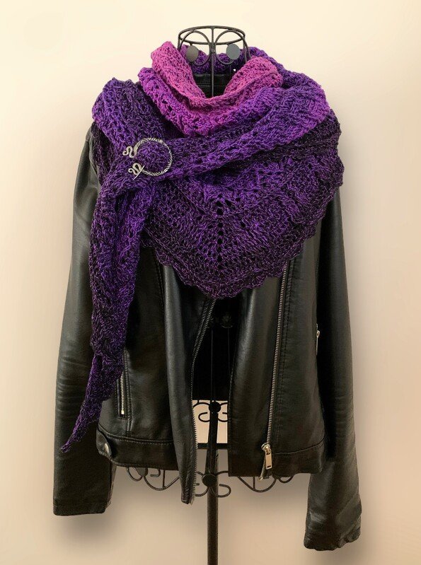 Purple to pink gradient crochet triangle shawl with openwork stitch, pinned with a decorative brooch on a black leather jacket.