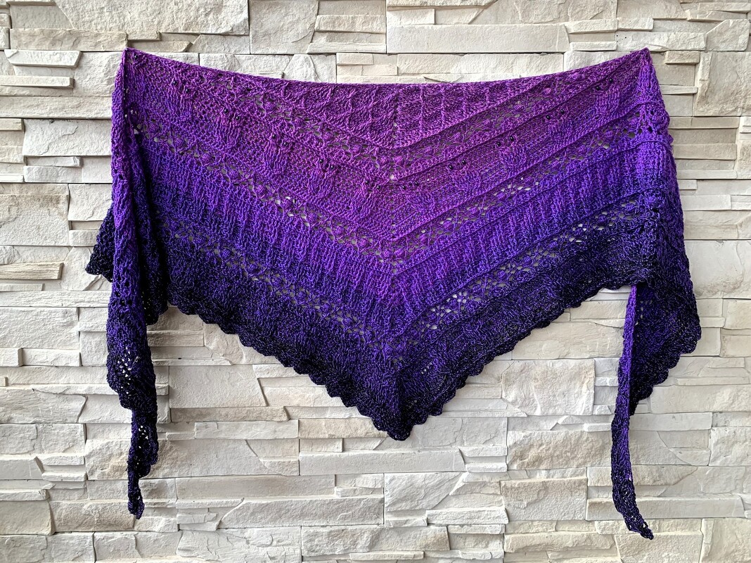 Purple ombre crocheted triangle shawl with lace pattern hanging on a light brick wall