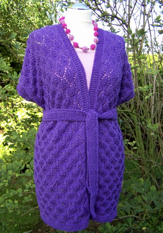 Lila gestrickte Damenjacke mit kurzen Ärmeln, Bindegürtel und Rauten-Ajourmuster auf einer Schneiderpuppe vor Pflanzen