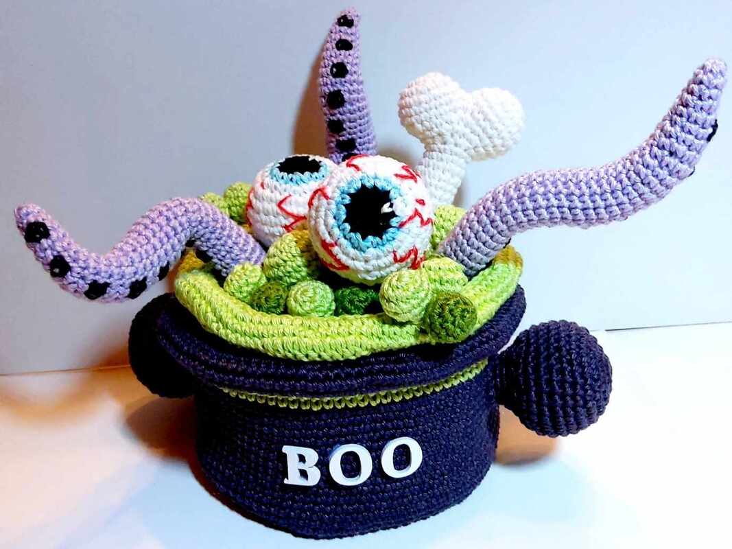 Amigurumi-Hexenkessel in Dunkelblau mit grüner Füllung, zwei weißen Augen, lilafarbenen Tentakeln, kleinem Knochen und 'BOO'-Schriftzug.
