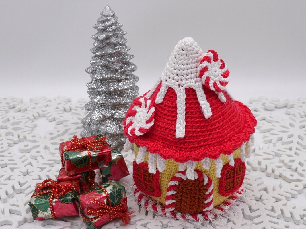 Gehäkeltes Lebkuchenhaus in Rot und Weiß mit Zuckerstangen-Akzenten, kleinen Geschenkpaketen und silbernem Baum auf weißem Schneeflocken-Untergrund