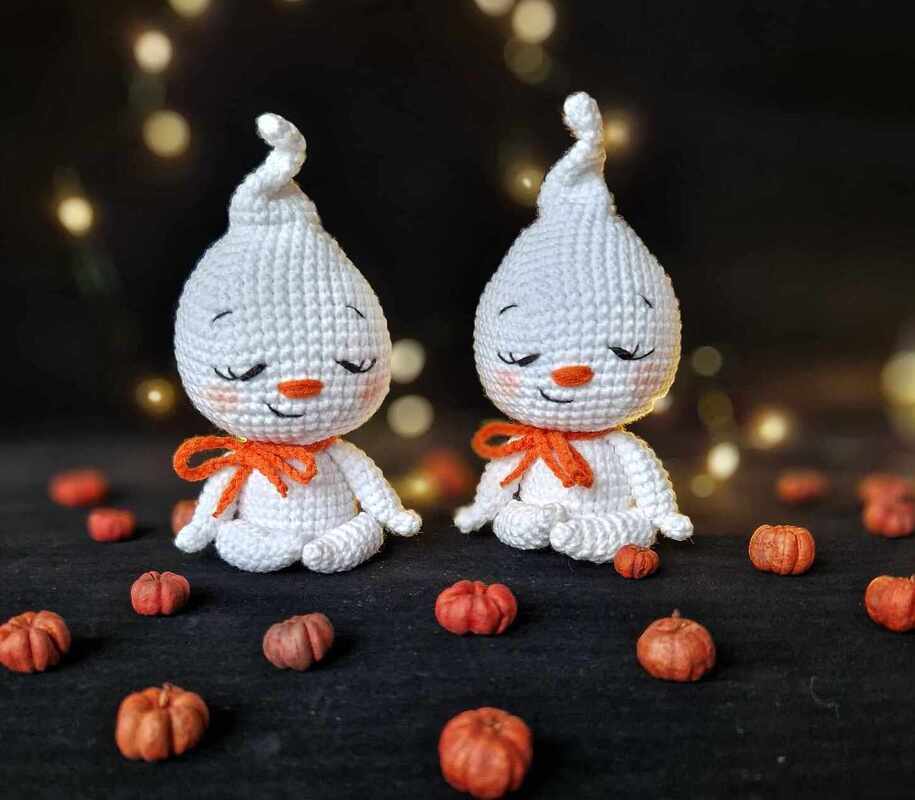 Two white crocheted ghost amigurumi with orange bows and small pumpkins on a dark surface